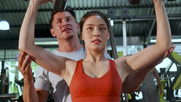 Fitness trainer and beginner sportswoman workout by lifting weight with barbell together in gym alt