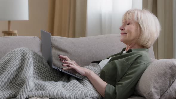 Old Senior Caucasian Gray Haired Female Sit on Couch Covered with Blanket Typing Laptop Write Email alt