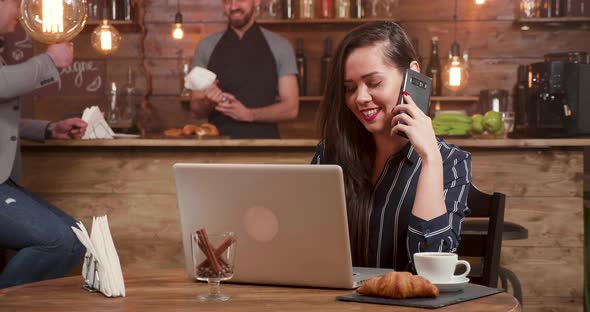 Young Pretty Lady Talking at Her Phone While Working at Her Laptop Remotely alt