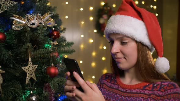 Portrait of Young Beautiful Woman with Santa Hat in Winter Sweater Typing Message on Mobile Phone