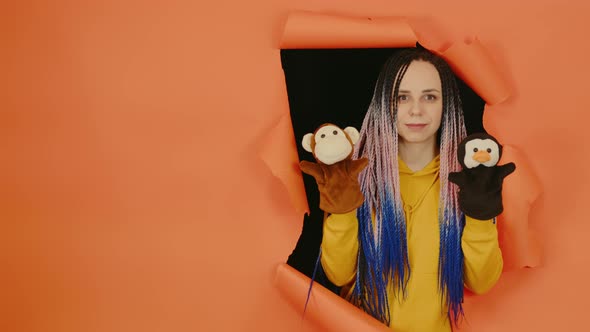 Young Woman with Soft Puppet Toys on Hands Looking Out of Hole of Orange Background alt