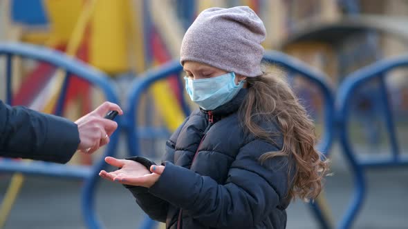 A girl in a medical mask. Her mother sprays disinfectant on her hands in the Playground