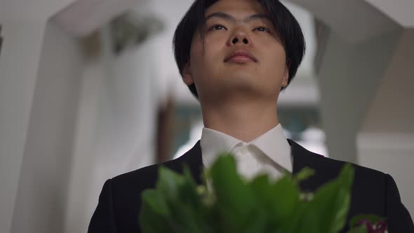 Closeup Portrait of Happy Young Asian Groom Standing with Bouquet Indoors Looking Away alt