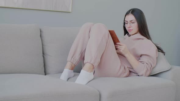 A young Caucasian woman wearing reading glasses reads a book on the sofa in a cozy home environment