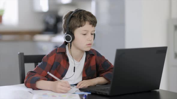 Boy listening to online class on laptop, writing in notebook