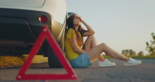 Young Upset Girl Sitting Next to Broken Car on Side of Road and Waiting Help alt