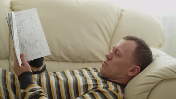 Portrait of a young man. Lying on the couch, reading a book. Moving camera