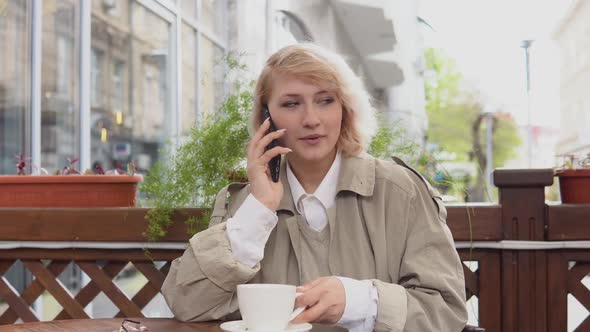Woman Talking on a Cell Phone with a Cup of Coffee at a Table on the Terrace alt