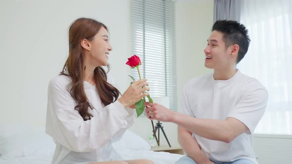Asian young man surprise and give rose flower to girlfriend in bedroom. alt