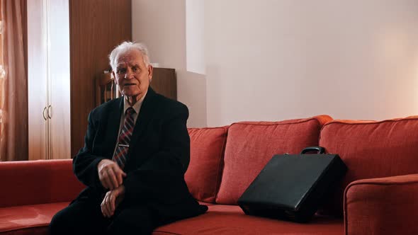 Elderly Grandfather - Grandfather Is Sitting on the Couch with a Suitcase and Checking the Time on alt