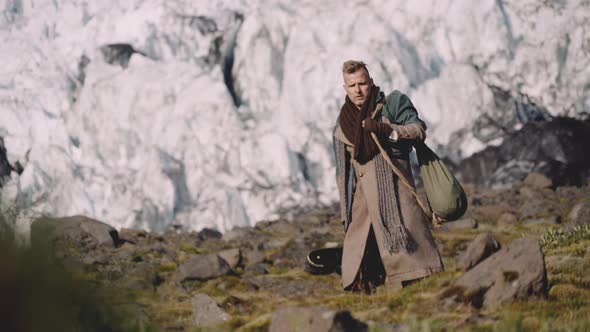 Man In Greatcoat Carrying Guitar Case And Bag Away From Glacier alt