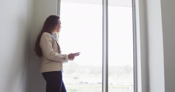 Biracial businesswoman standing using smartphone by window in modern interiors alt