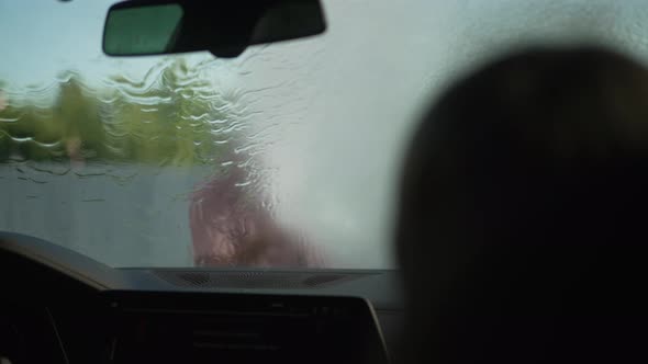 Car Windshield with Water Flowing in Slow Motion and Blurred Man Waving As Silhouette of Boy alt