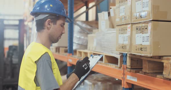 Logistics worker wearing a helmet working in a large warehouse checking inventory alt