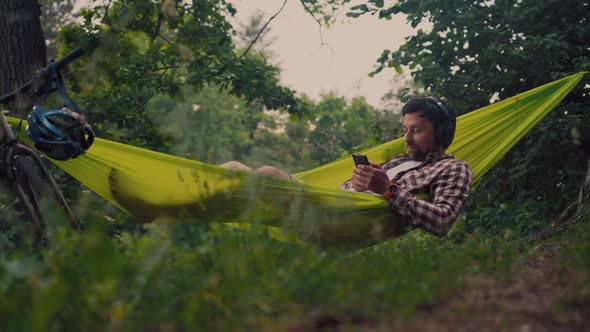 Man on Bicycle Trip at Camping By Lake is Relaxing in Green Hammock While Listening to Music alt
