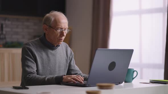 Elderly Man with Glasses is Working with Laptop at Home Surfing Internet and Looking for Information alt