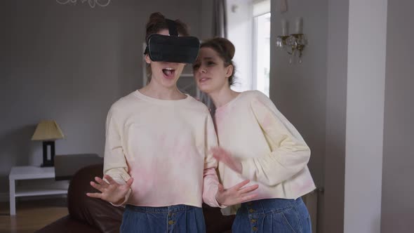Happy Absorbed Young Woman in VR Headset Enjoying Augmented Reality As Curios Twin Sister Asking to alt