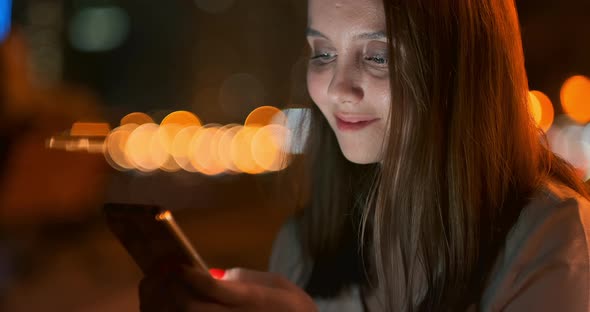 A Young Girl on the Background of the Night City Looks Into the Smartphone Screen alt