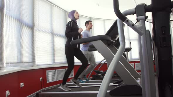 Female Athlete in a Hijab on a Running Simulator and a Man in a Gym Family alt
