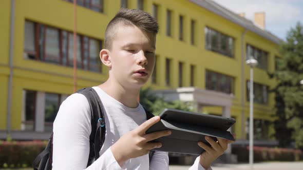 A Caucasian Teenage Boy Talks to the Camera and Holds a Tablet  Closeup  a School in Background