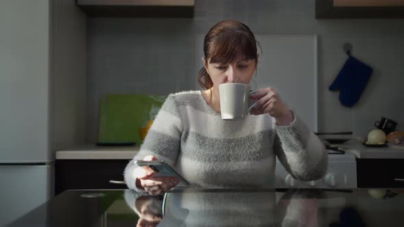 Pensive Woman Sitting in the Kitchen at Home with a Cup of Tea and Uses the Phone