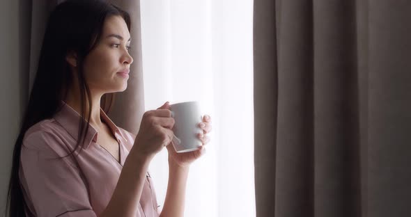 Woman Stands Near the Window with the Curtains Open Drinking Tea From a Mug