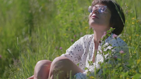 Woman in Colorful Sunglasses Enjoys Sunlight and Flower Fragrance on Grass Field alt