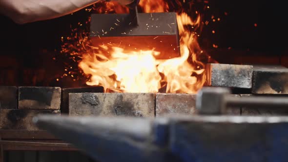 Worker Throws Coals in Fire with Hammer on Anvil on the Foreground.