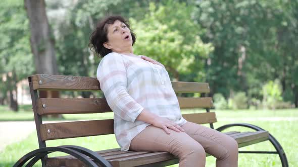 Ill Caucasian Senior Woman Having Heart Attack Sitting on Bench in Summer Park As People Passing By alt