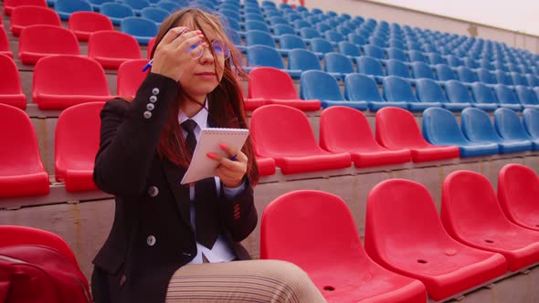 Young Woman in Glasses with Notepad Pen Sitting on Stadium Bleachers Alone alt