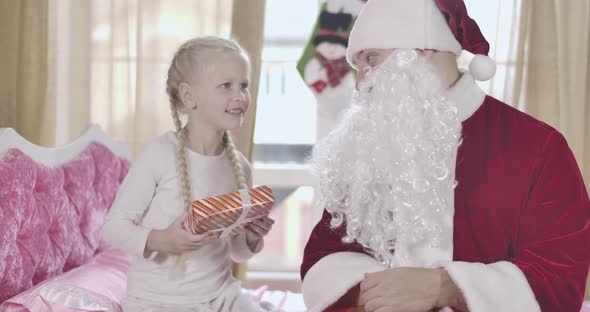 Portrait of Cute Blond Caucasian Girl with Pigtails Rubbing Hands As Santa Claus Giving Her alt