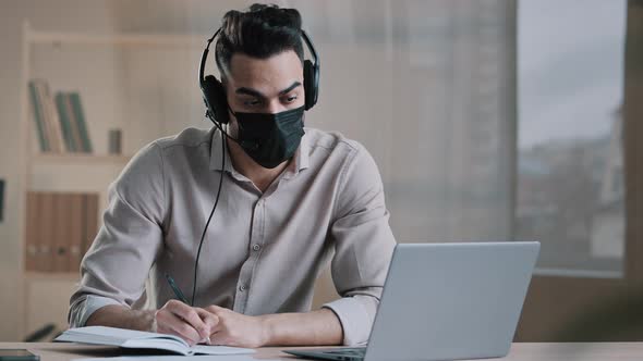 Young Arab Man Freelancer Male Writer in Medical Mask Wear Headphones Sit Home Office During alt