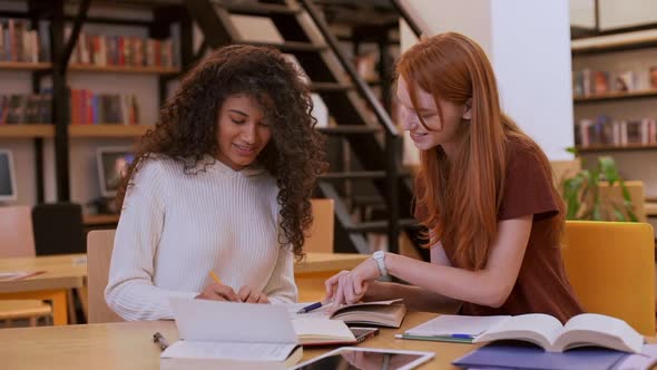 Two Teenage Girls Sitting at Library While Boy in Shirt and Glasses Coming to Them Discussing Book alt