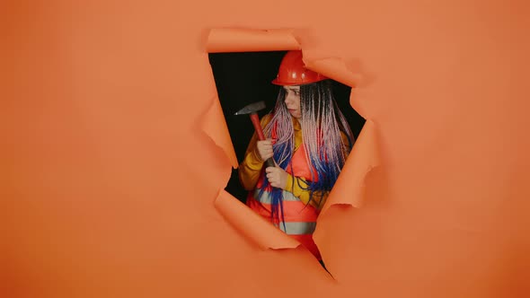 Shocked Female Construction Worker in Overalls with Hammer Looking Out of Hole of Orange Background alt