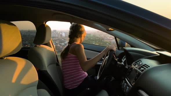 Young woman standing near her car enjoying warm sunset view. Girl traveler leaning on vehicle  alt