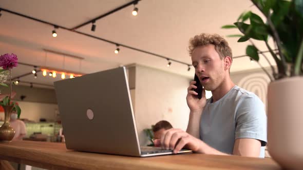 Young man working on laptop indoors. He talks to someone on the phone alt