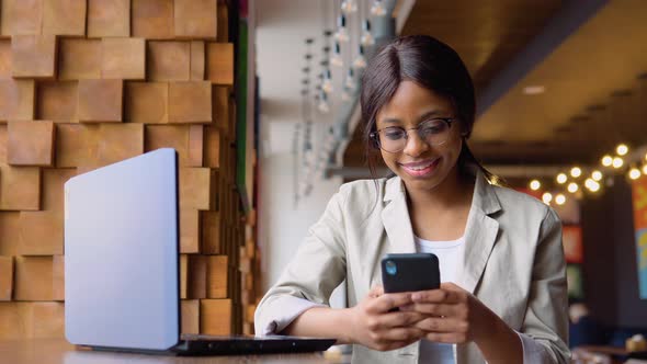 Young Indian Woman in Glasses Using Phone While Sitting in Cafe with Laptop alt