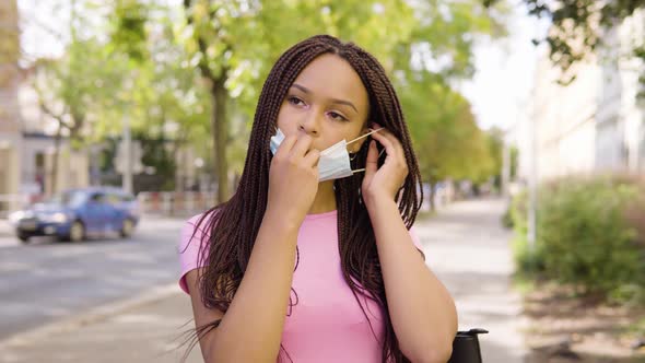 A Young Black Woman Puts on a Face Mask and Looks at the Camera in the Street in an Urban Area alt
