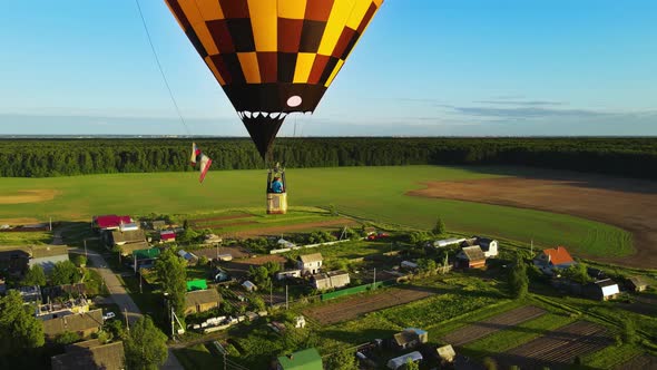 Two aeronauts in gondola fly balloon. A yellow balloon over village at sunset. alt