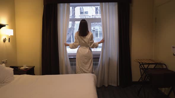 A Woman in a Hotel Room Opens the Curtains By the Window Early in the Morning