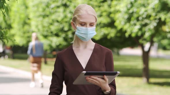 A Woman in a Medical Mask Stands in the Middle of the Street and Scrolls a Tablet