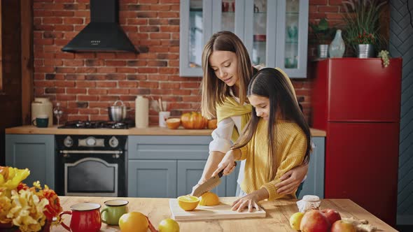Young Mum Teaching Little Daughter How to Cut Orange with Knife Standing By Table in Modern Kitchen alt