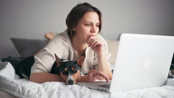 A Middleaged Woman Is Working on Her Laptop While Lying on the Bed with Her Dog alt