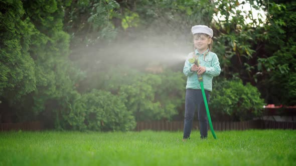 A Small Child is Watering the Plants Near the House with a Hose