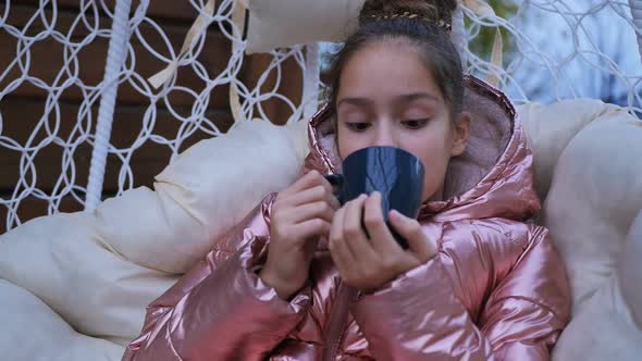 Cute Girl in a Jacket is Happy to Drink Tea From a Glass in a Street Chair alt