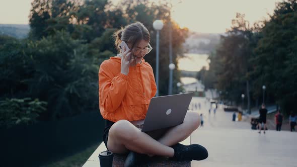 A Hardworking and Optimistic Girl Talks on the Phone Smiles Broadly While Typing on the Laptop alt