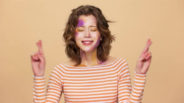 Portrait of Concentrated Woman with Multicolored Makeup Stains on Face Holding Fingers Crossed As alt