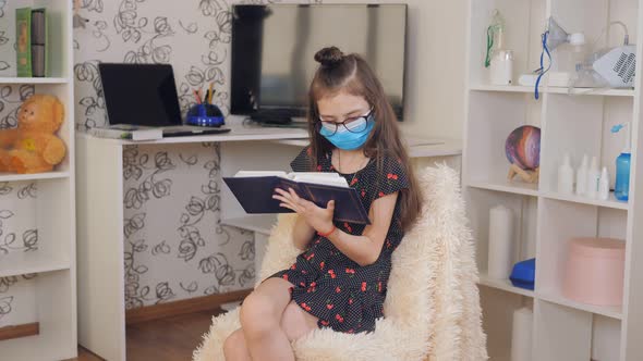 A Girl with Glasses and a Medical Mask Sitting in the Room on a Chair and Looking at the Pictures in