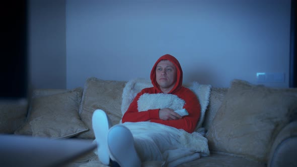 Man in a Hooded Sweatshirt Sitting on the Couch at Night with His Feet in Front of Him Watching TV alt