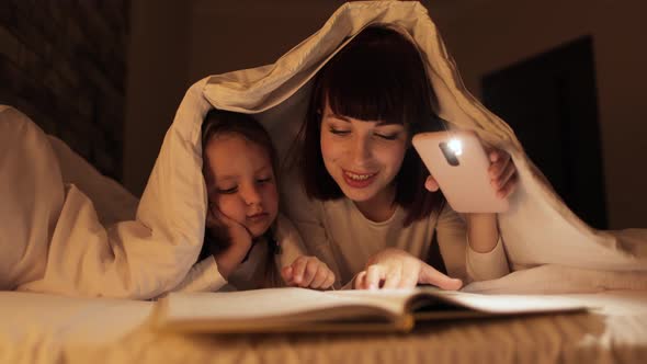 Young Affectionate Mother and Child Daughter Reading Book Using a Flashlight Before Going to Bed alt